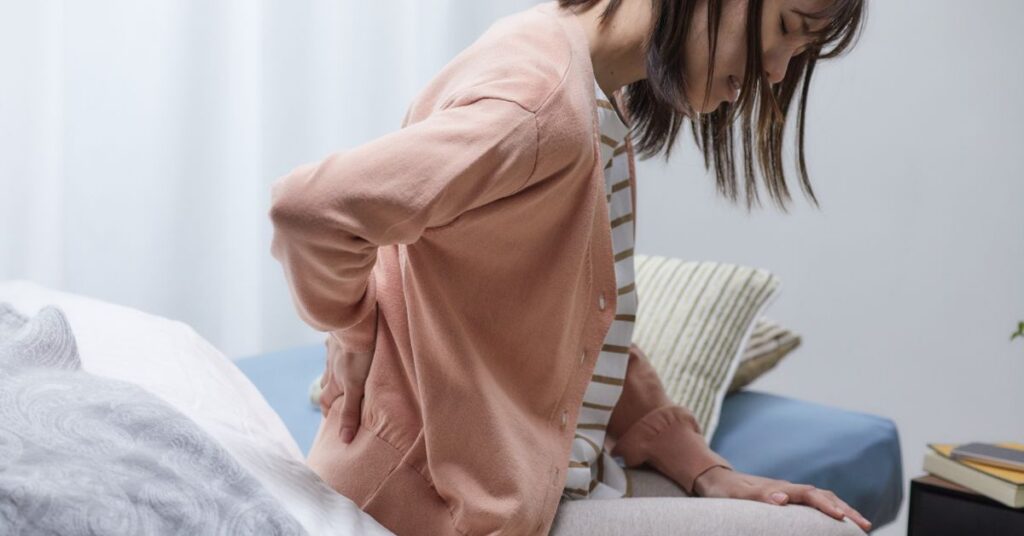 Woman sitting on a bed holding her lower back in severe pain from a delayed car accident injury.